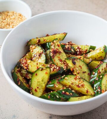 A white bowl of vegan cucumber salad covered in chili next to a white pot of sesame seeds