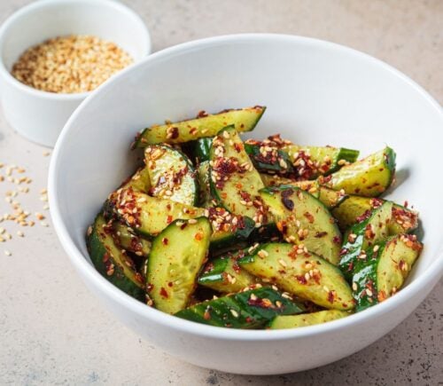 A white bowl of vegan cucumber salad covered in chili next to a white pot of sesame seeds