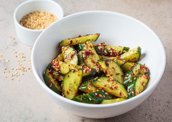 A white bowl of vegan cucumber salad covered in chili next to a white pot of sesame seeds