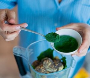 Photo shows someone spooning dark green spirulina into a blender to make smoothies