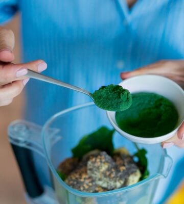 Photo shows someone spooning dark green spirulina into a blender to make smoothies