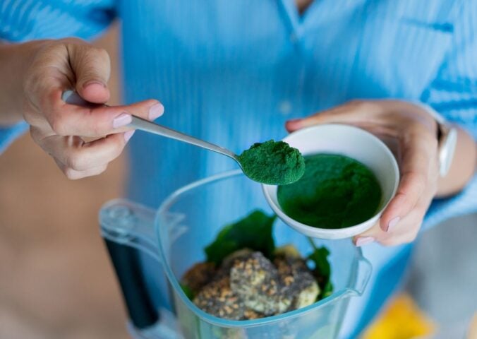 Photo shows someone spooning dark green spirulina into a blender to make smoothies