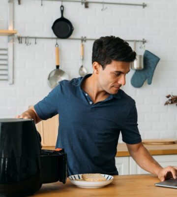 Photo shows a man using his laptop and operating an air fryer while standing at a kitchen island