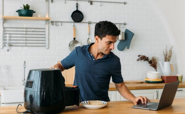 Photo shows a man using his laptop and operating an air fryer while standing at a kitchen island