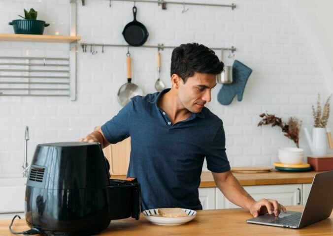 Photo shows a man using his laptop and operating an air fryer while standing at a kitchen island