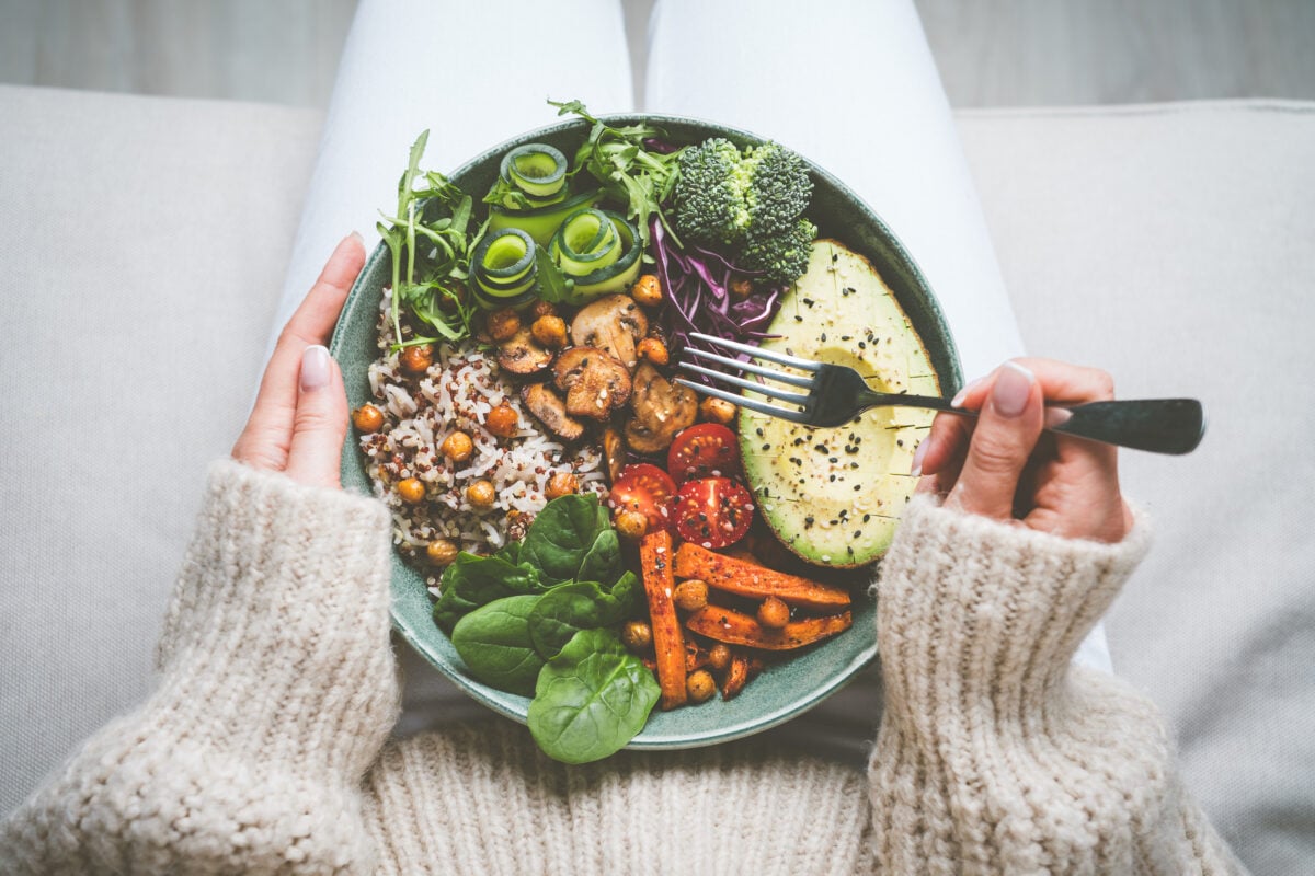 A person in a cosy jumper eating a bowl of plant-based whole foods including avocado, rice, and other vegetables