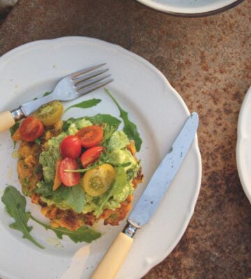 a picture of a corn fritter topped with avocado smash, arugula, and tomato