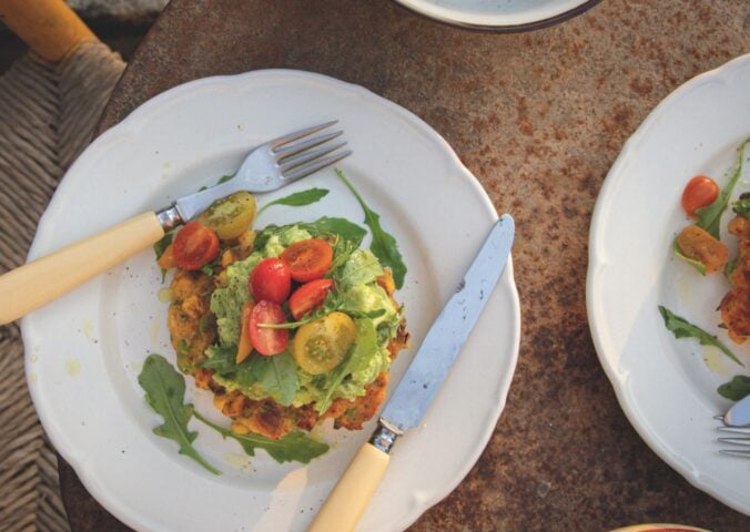 a picture of a corn fritter topped with avocado smash, arugula, and tomato