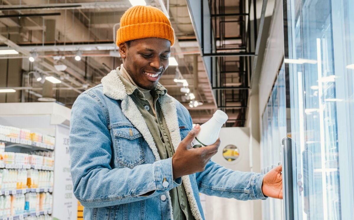 Photo shows a young man reading the packaging on a drink while standing in the refrigerator aisle of a supermarket