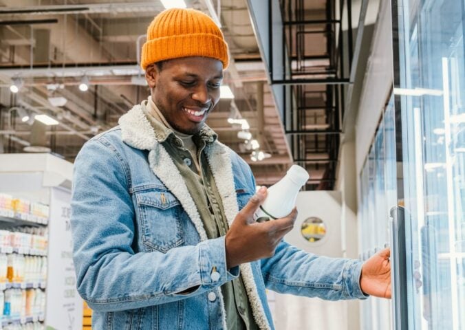 Photo shows a young man reading the packaging on a drink while standing in the refrigerator aisle of a supermarket