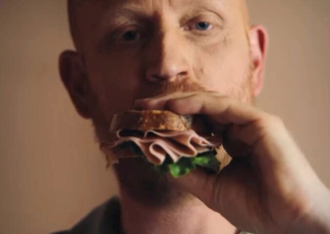 Image is a still taken from La Vie's new TV advertisement, it shows a man taking a bite of a vegan ham sandwich