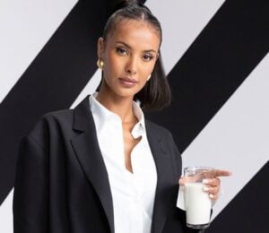 Maya Jama standing in front of a white and black striped background in a suit holding a glass of milk