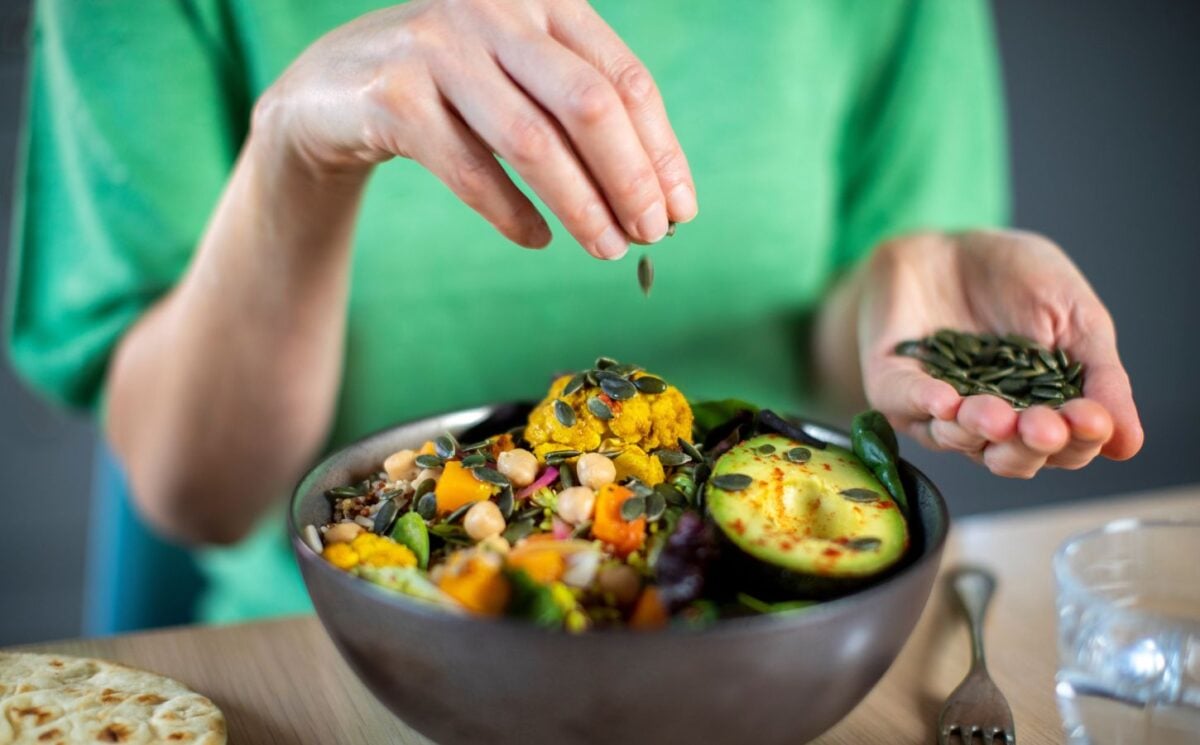 Photo shows someone sprinkling nuts and seeds onto a bowl of colorful plant-based food