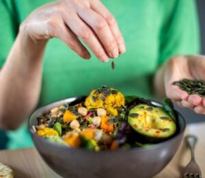 Photo shows someone sprinkling nuts and seeds onto a bowl of colorful plant-based food