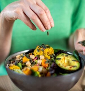 Photo shows someone sprinkling nuts and seeds onto a bowl of colorful plant-based food