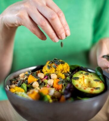 Photo shows someone sprinkling nuts and seeds onto a bowl of colorful plant-based food