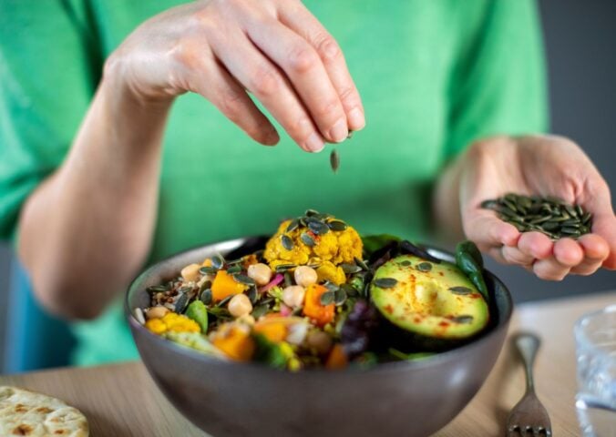 Photo shows someone sprinkling nuts and seeds onto a bowl of colorful plant-based food