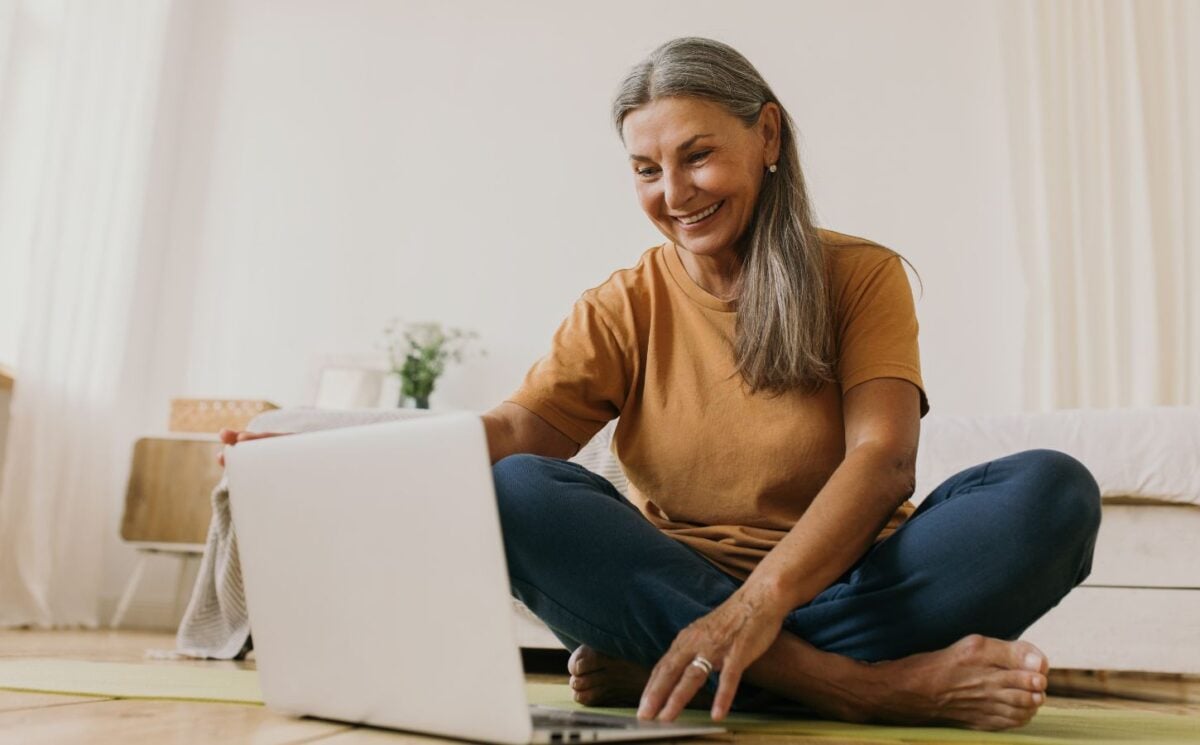 A woman sat on the floor smiling and looking at her laptop