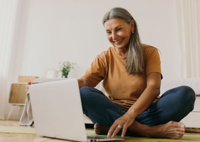 A woman sat on the floor smiling and looking at her laptop