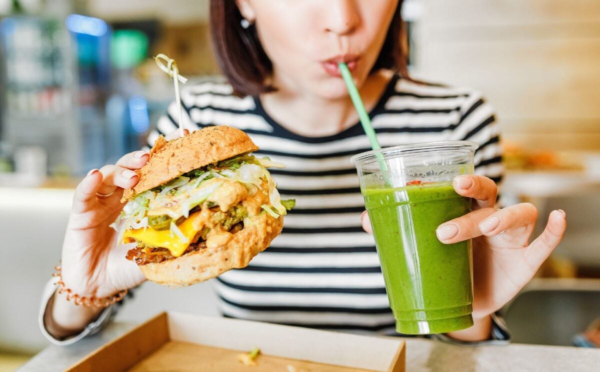 A woman drinking a green smoothie and eating a vegan burger at a brightly-lit vegan restaurant