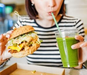 A woman drinking a green smoothie and eating a vegan burger at a brightly-lit vegan restaurant