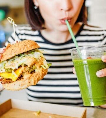 A woman drinking a green smoothie and eating a vegan burger at a brightly-lit vegan restaurant