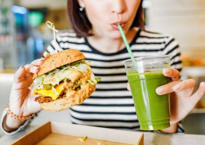 A woman drinking a green smoothie and eating a vegan burger at a brightly-lit vegan restaurant