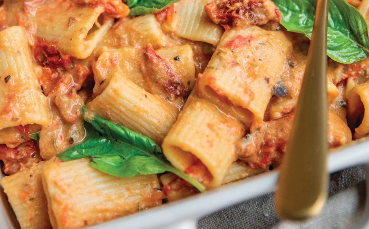 a baked hummus pasta dish with hummus, sun-dried tomatoes, tomato, rigatoni, and basil