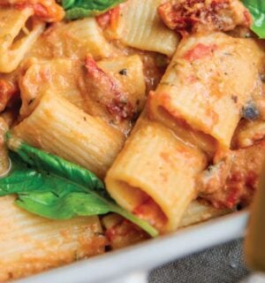 a baked hummus pasta dish with hummus, sun-dried tomatoes, tomato, rigatoni, and basil