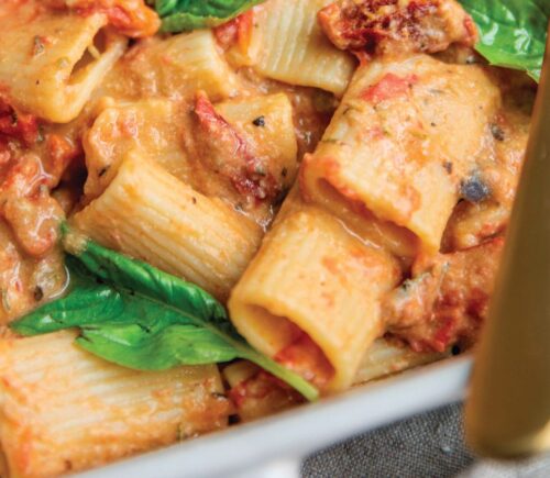 a baked hummus pasta dish with hummus, sun-dried tomatoes, tomato, rigatoni, and basil