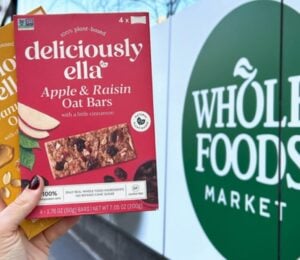 Photo shows someone's hand as they hold up the two flavors of Deliciously Ella's oat bars in front of a Whole Foods Market store