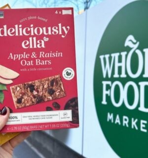 Photo shows someone's hand as they hold up the two flavors of Deliciously Ella's oat bars in front of a Whole Foods Market store