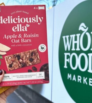 Photo shows someone's hand as they hold up the two flavors of Deliciously Ella's oat bars in front of a Whole Foods Market store