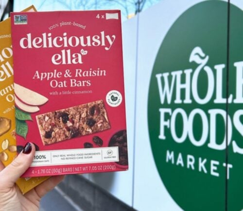 Photo shows someone's hand as they hold up the two flavors of Deliciously Ella's oat bars in front of a Whole Foods Market store