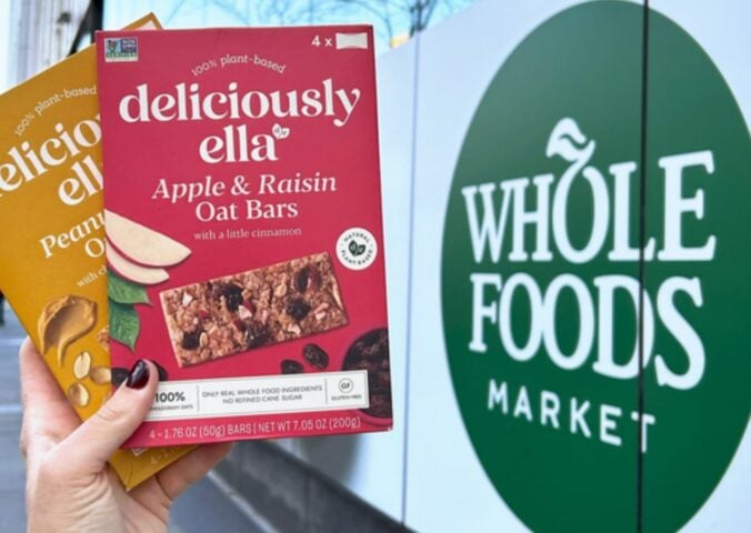 Photo shows someone's hand as they hold up the two flavors of Deliciously Ella's oat bars in front of a Whole Foods Market store