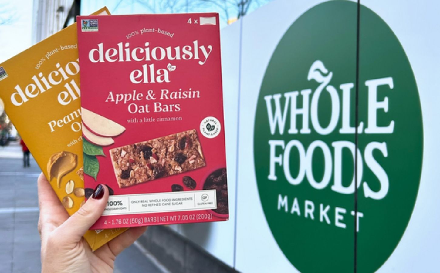 Photo shows someone's hand as they hold up the two flavors of Deliciously Ella's oat bars in front of a Whole Foods Market store