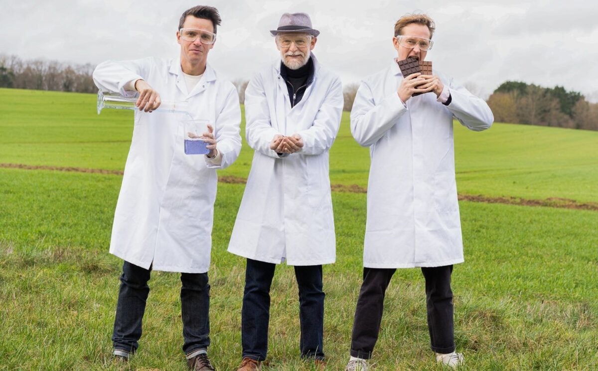 Photo shows Nukoko founders Kit Tomlinson, Ross Newton, and David Salt standing in a field, wearing lab coats, and holding up their proprietary bean-based chocolate alternative