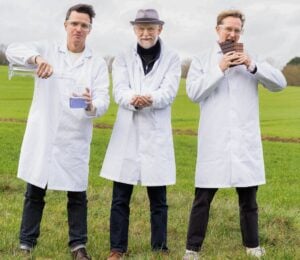 Photo shows Nukoko founders Kit Tomlinson, Ross Newton, and David Salt standing in a field, wearing lab coats, and holding up their proprietary bean-based chocolate alternative