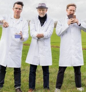 Photo shows Nukoko founders Kit Tomlinson, Ross Newton, and David Salt standing in a field, wearing lab coats, and holding up their proprietary bean-based chocolate alternative