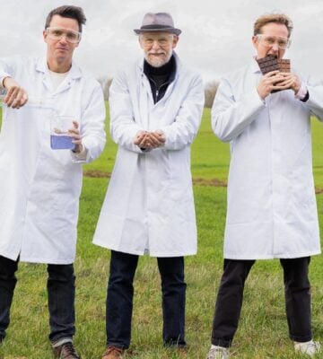 Photo shows Nukoko founders Kit Tomlinson, Ross Newton, and David Salt standing in a field, wearing lab coats, and holding up their proprietary bean-based chocolate alternative