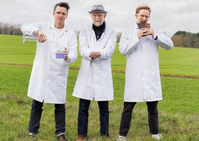 Photo shows Nukoko founders Kit Tomlinson, Ross Newton, and David Salt standing in a field, wearing lab coats, and holding up their proprietary bean-based chocolate alternative