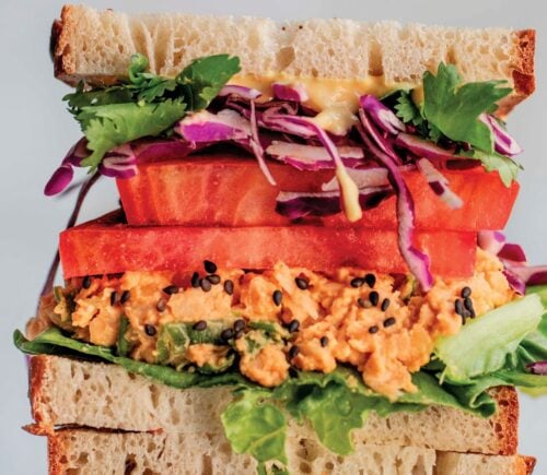 a picture of stacked peanut miso chickpea salad sandwiches with sesame, tomato, chickpea spread, and greens