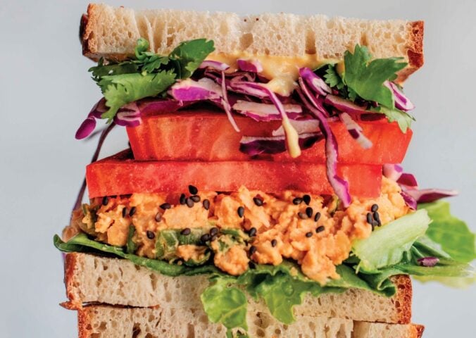 a picture of stacked peanut miso chickpea salad sandwiches with sesame, tomato, chickpea spread, and greens