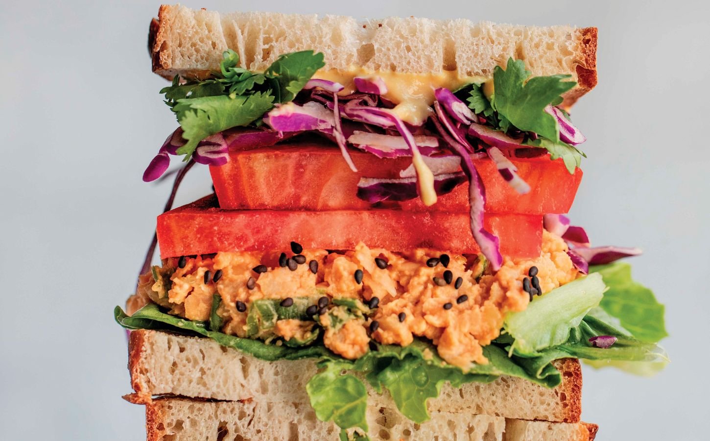 a picture of stacked peanut miso chickpea salad sandwiches with sesame, tomato, chickpea spread, and greens