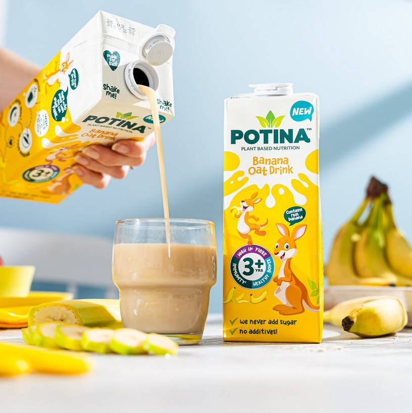 Photo shows someone pouring a glass of Banana Oat flavor Potina on a table with another carton and some bananas