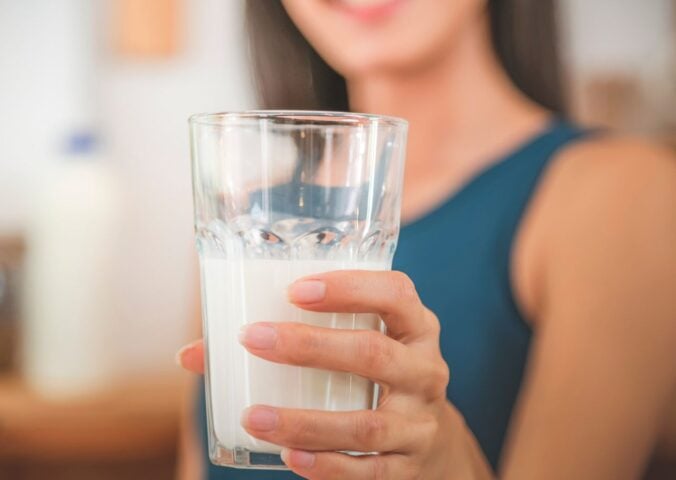 Photo shows a woman holding up a glass half full of milk