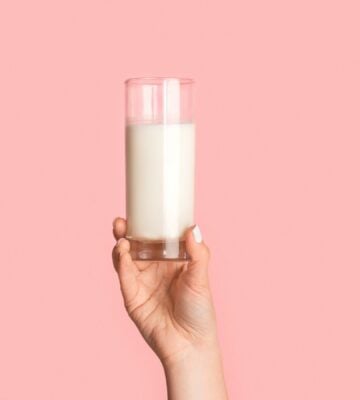 Photo shows a woman's hand as she holds up a glass of milk against a pale pink background