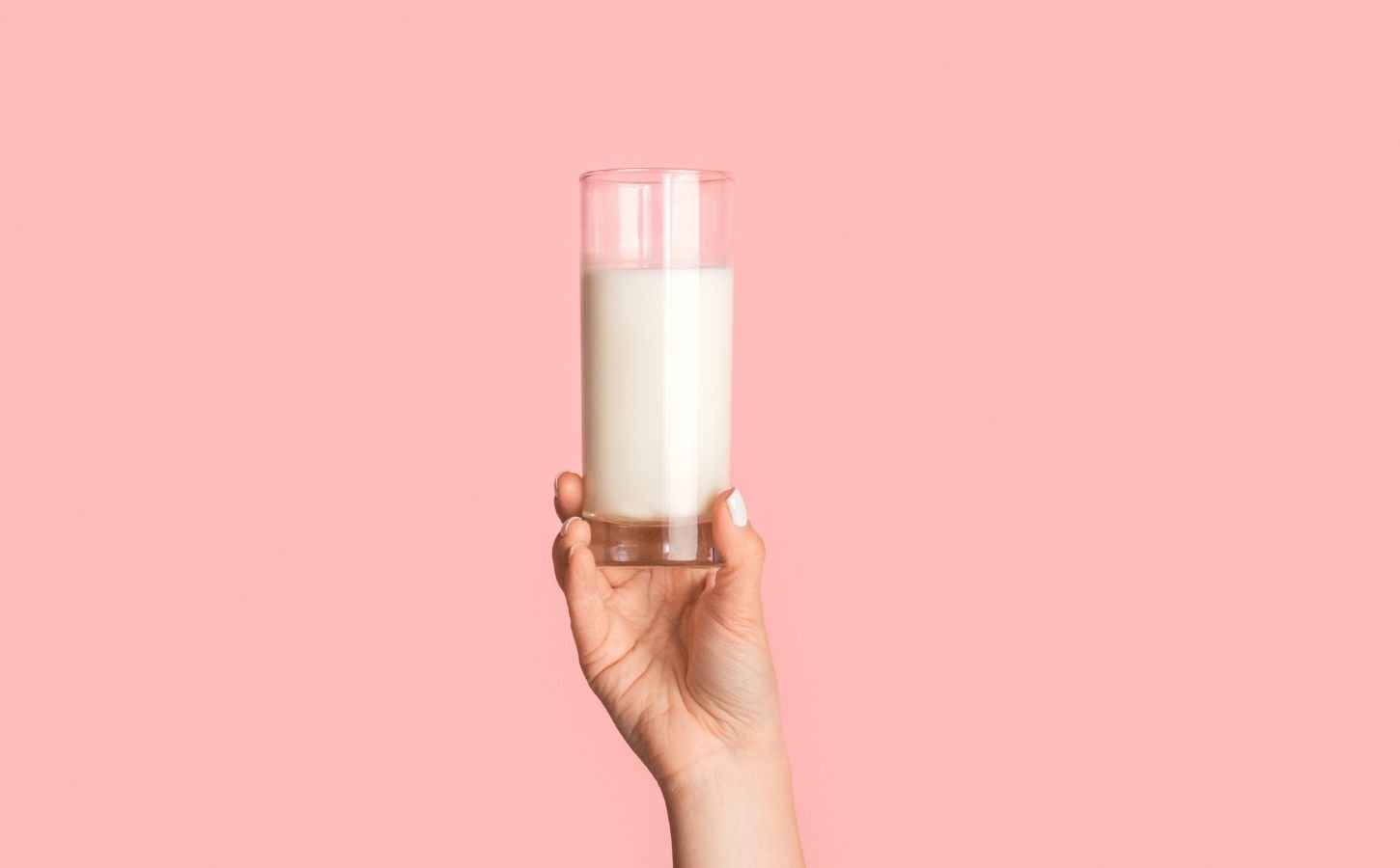 Photo shows a woman's hand as she holds up a glass of milk against a pale pink background