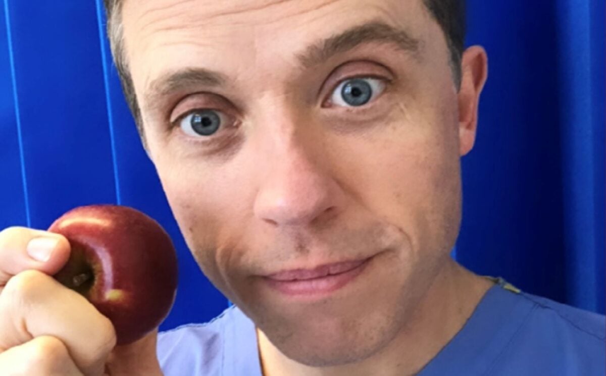 Renowned plant-based doctor Dr Alan Desmond holding an apple in front of some blue curtains at a hospital