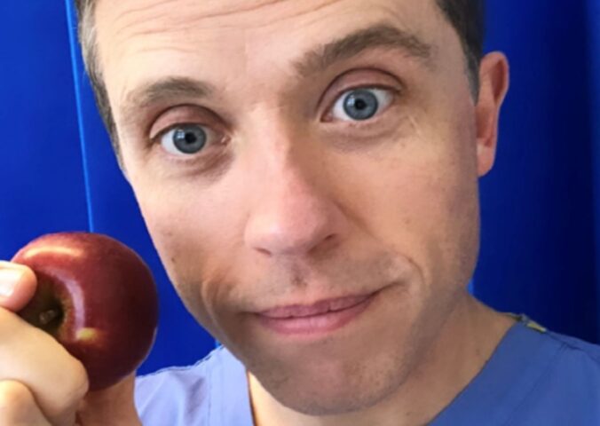 Renowned plant-based doctor Dr Alan Desmond holding an apple in front of some blue curtains at a hospital